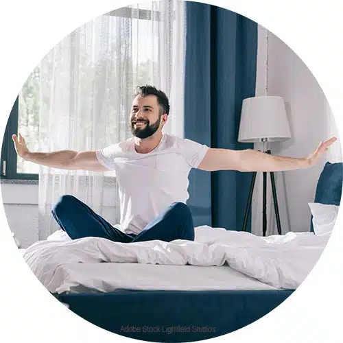 un home barbu assis sur un lit écarte les bras
